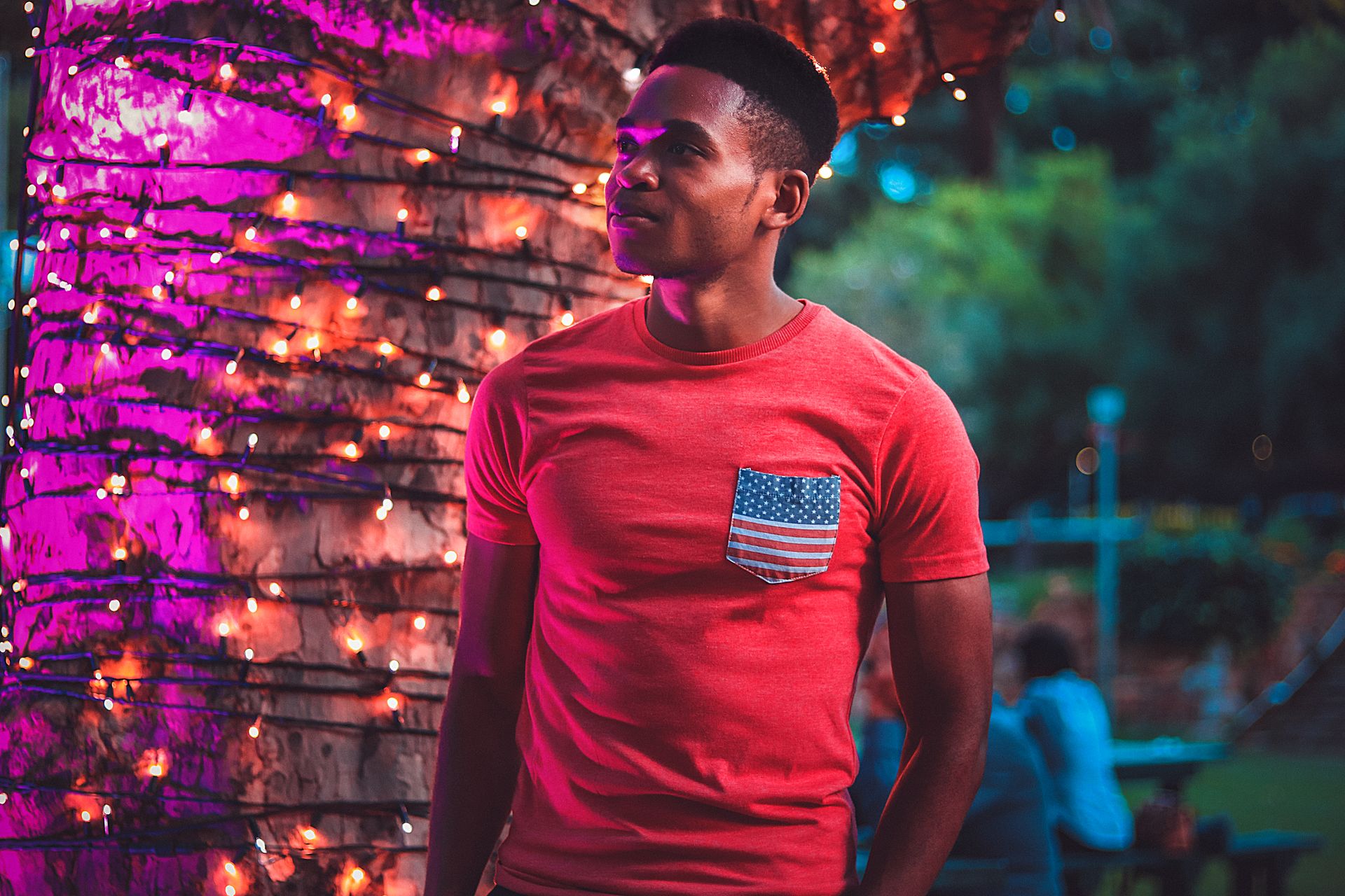 Portrait of a young man in a red t-shirt standing amidst colorful festive lights outdoors at night.
