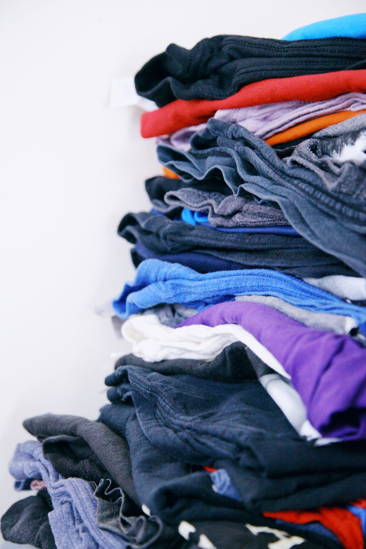 A tall, untidy stack of folded T-shirts in mixed colours