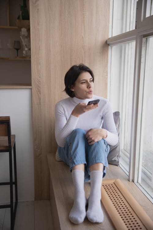 Young female in casual clothes without shoes sitting on windowsill with legs and looking at window while chatting on mobile phone in modern apartment