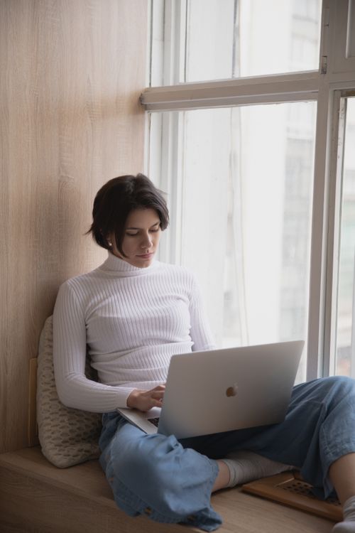 Free Focused woman in casual wear works remotely on laptop by a window. Stock Photo