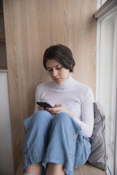 一名年轻女子坐在窗前，在室内用智能手机享受片刻宁静