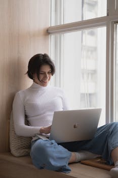 Woman in casual attire working on a laptop by a sunny window, embodying modern remote work lifestyle.