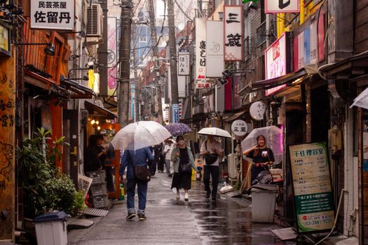 People with umbrellas stroll through a vibrant, rainy alleyway in Tokyo, surrounded by shops and signs.