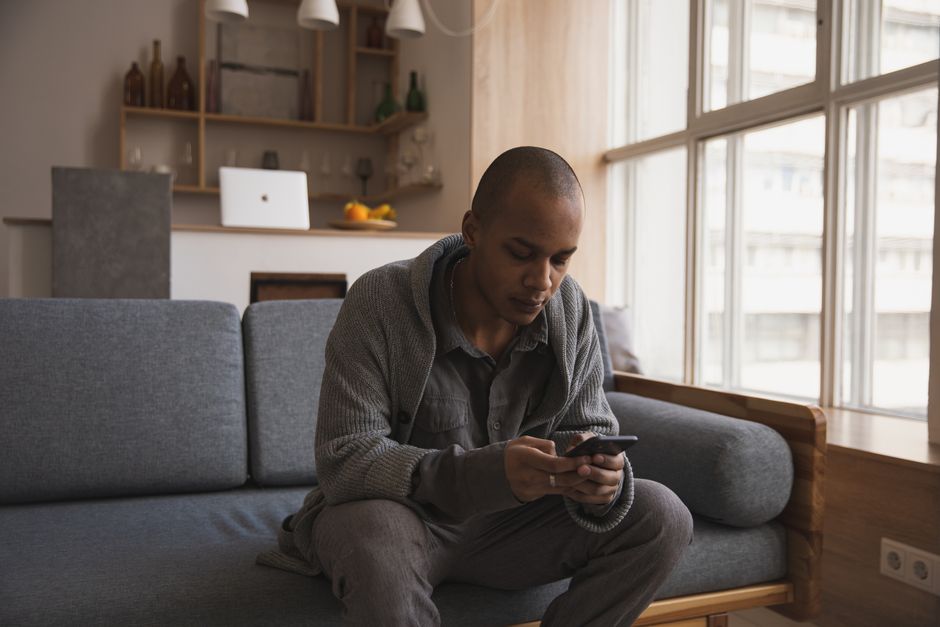 Dark-skinned Nigerian man in a grey hoodie sitting at a desk in a small London flat, looking at his phone with a relieved expression, a damp UK winter day visible through the window behind him, photorealistic