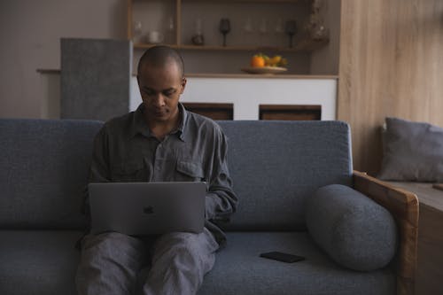 Serious young African American man in casual outfit using netbook while sitting on sofa in modern living room and chatting on social media