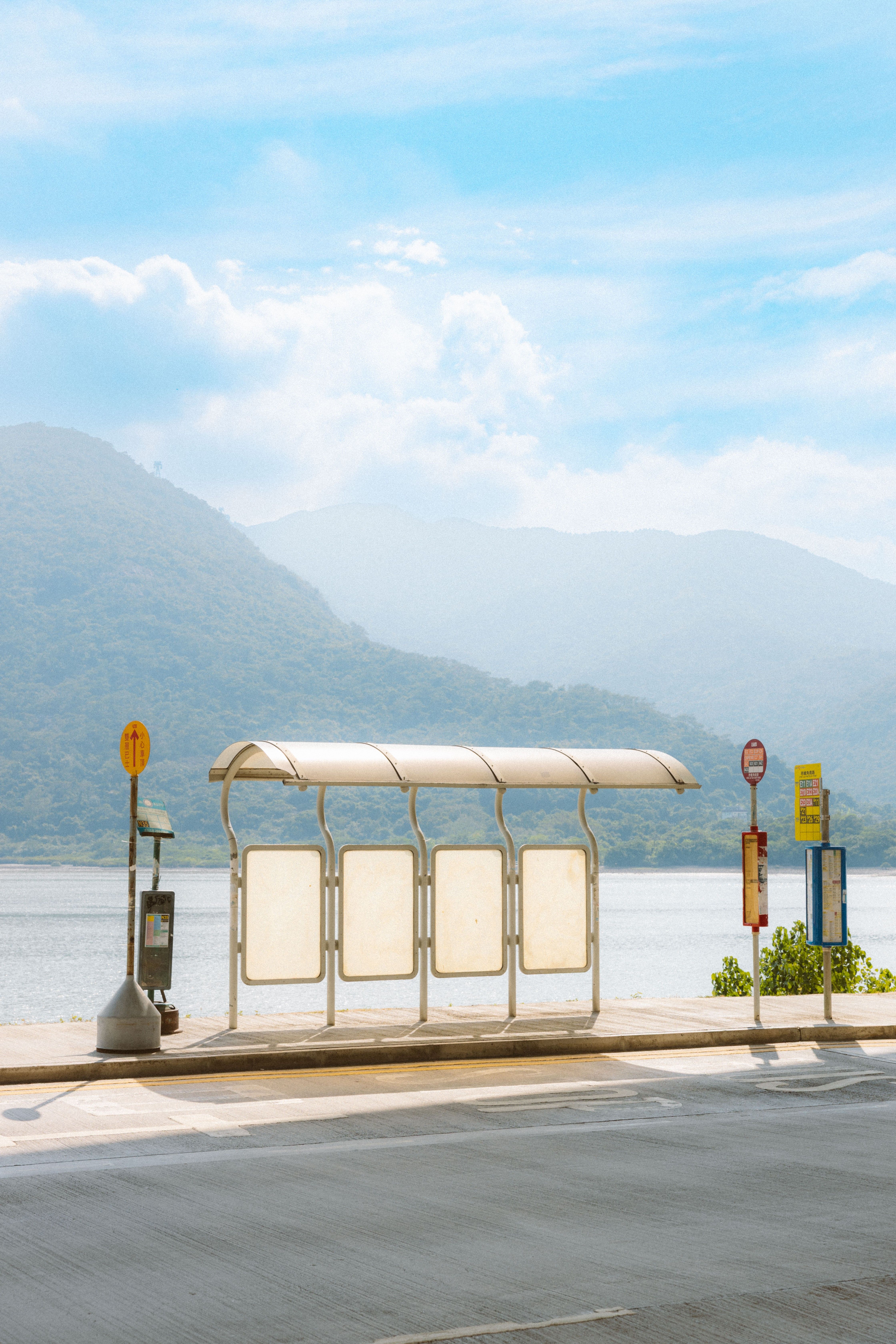 Empty bus stop by the water with mountains in the background under a bright blue sky.