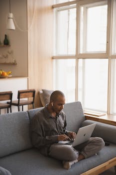 Man sitting on couch working on a laptop in a cozy home setting, embodying remote work lifestyle.