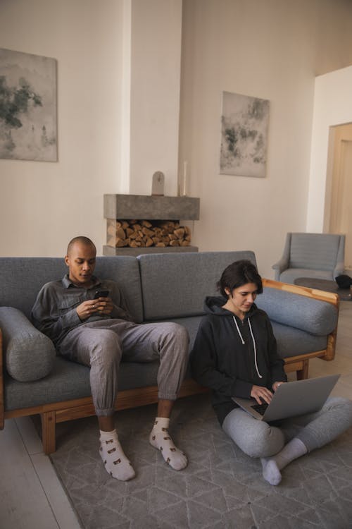 From above of African American man in casual wear using smartphone and sitting on sofa and woman sitting with crossed legs on carpet near and typing on laptop at home