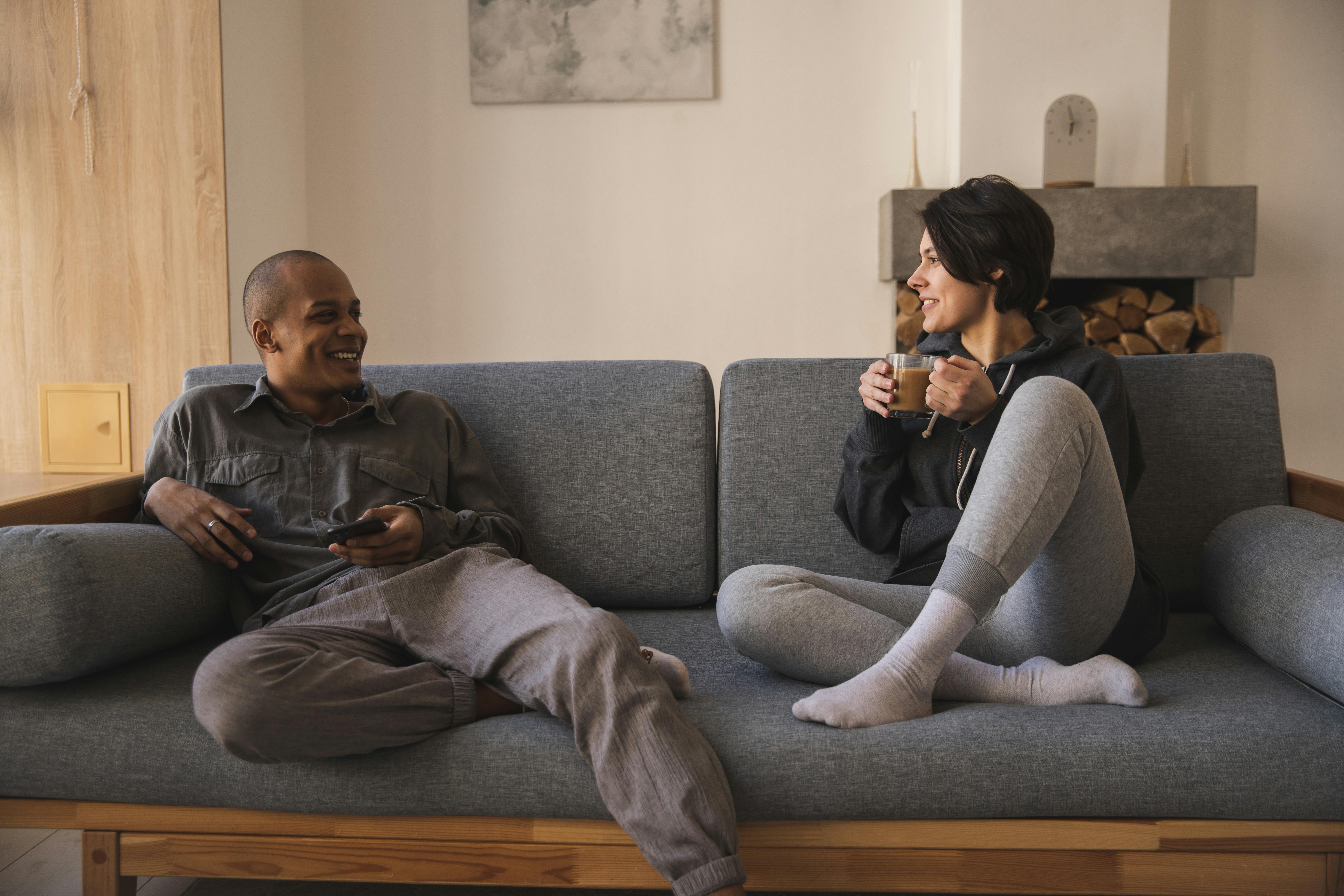 A couple is sitting on a blue couch having a deep conversation. The man is black and seated on the left, casually lounging on the couch. The woman is wearing grey sweatpants and holding a cup of coffee.