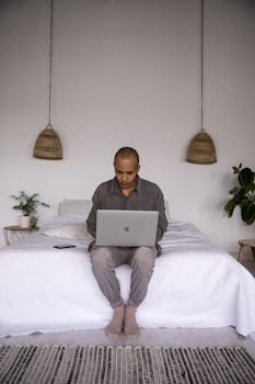 Man sitting on a bed, working on a laptop in a minimalist bedroom.