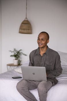 Smiling man sitting on bed working remotely with a laptop in a cozy bedroom setting.