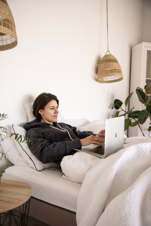 Free A woman comfortably works from bed using a laptop, surrounded by cozy bedroom decor. Stock Photo