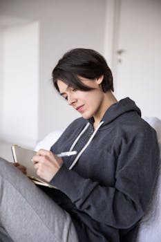A woman in a hoodie sits indoors writing in her journal with focus.