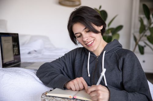 Free A woman in a hoodie sits on bed writing in a notebook, capturing a candid moment. Stock Photo