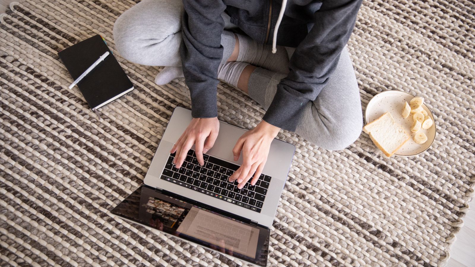 A person sitting cross-legged on the floor checking a laptop with breakfast nearby