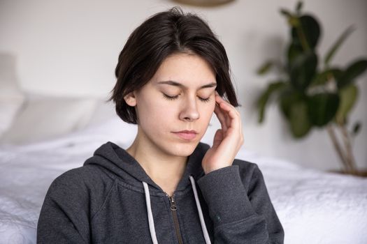 一位年轻女子闭着眼睛，穿着连帽衫，若有所思地坐在室内，仿佛在沉思或冥想