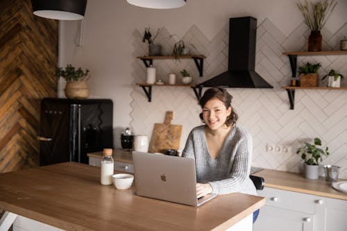 Cheerful female in casual clothes sitting at table with laptop and bottle of milk while browsing internet on laptop during free time at home and smiling looking at camera