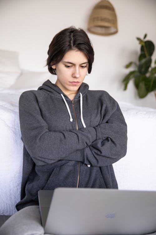 Free Serious female student in gray hoodie sitting with crossed arms on floor near bed and watching laptop while staying at home during online lesson Stock Photo