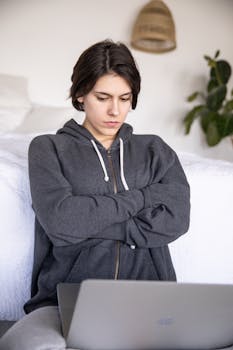 一名穿着灰色连帽衫的严肃女学生，在线上课期间呆在家里，双臂交叉坐在床边地板上看着笔记本电脑