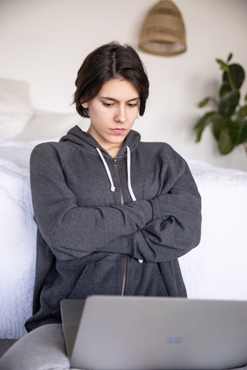 Serious female student in gray hoodie sitting with crossed arms on floor near bed and watching laptop while staying at home during online lesson