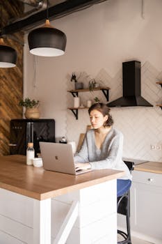 Young woman on a laptop, working remotely in a modern kitchen setting.