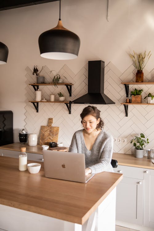 Focused woman in casual outfit sitting at counter with milk bottle in modern kitchen and browsing netbook while working on remote project at home