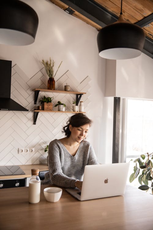 Free Cheerful young woman using laptop in kitchen Stock Photo