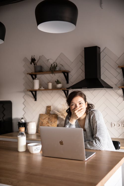 Free Young female in gray sweater covering mouth with hand while sitting at wooden table with laptop and milk bottle and having rest watching video while looking away Stock Photo