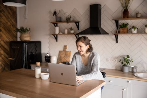 Free Young female in gray sweater laughing while sitting at wooden table with laptop and milk bottle and having rest watching video during free time Stock Photo