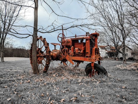 An old rusty tractor standing idle in a rural countryside farm setting, surrounded by leafless trees.