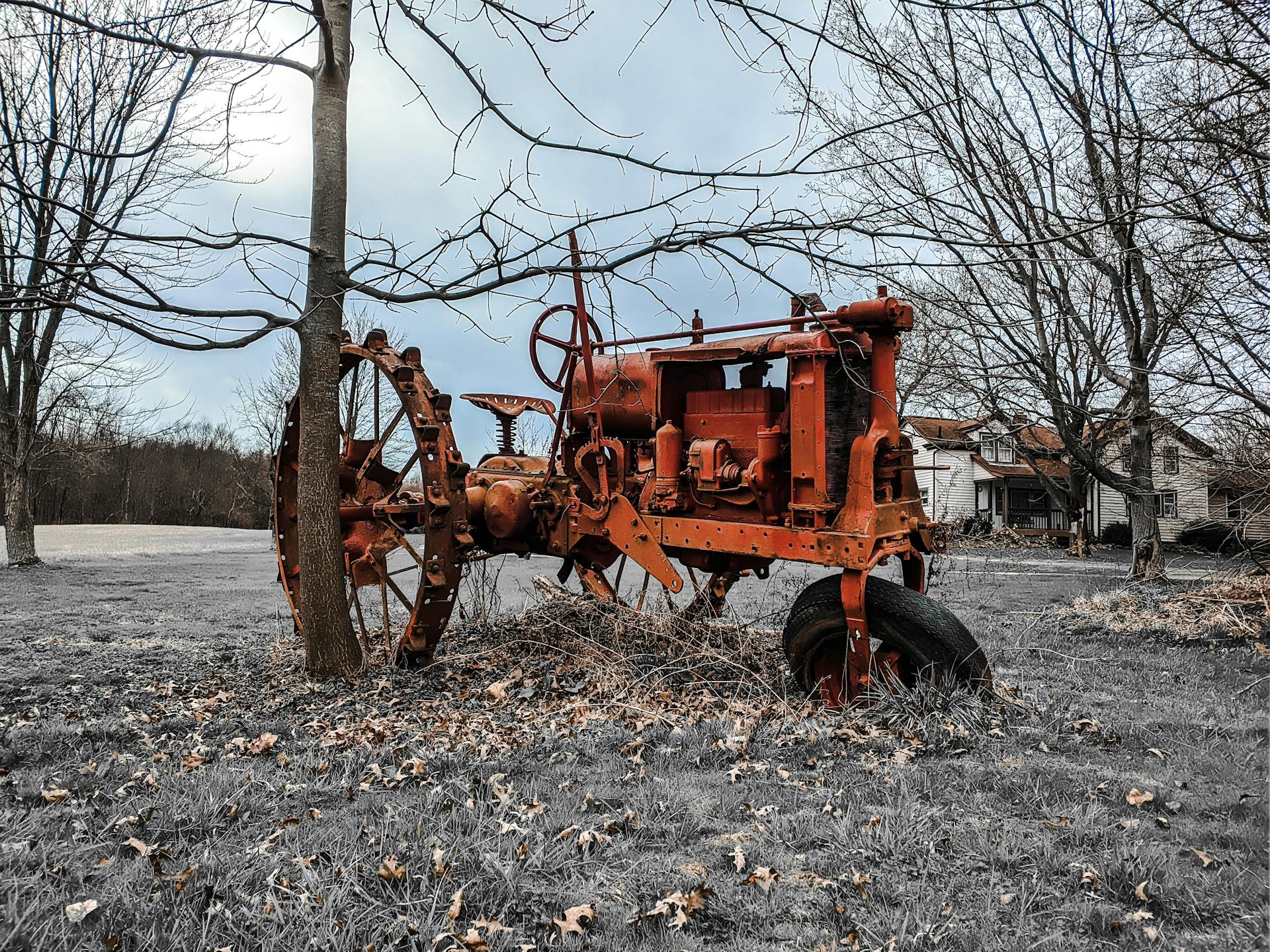 An old rusty tractor standing idle in a rural countryside farm setting, surrounded by leafless trees.