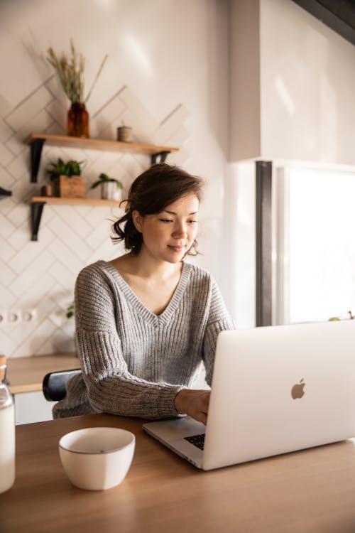 Young female in gray sweater sitting at wooden desk with laptop and bottle of milk near white bowl while browsing internet on laptop during free time at home