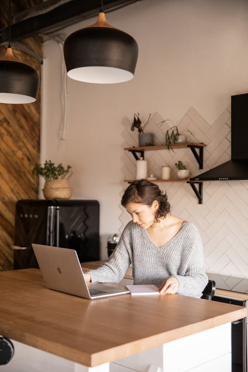 Young businesswoman in gray sweater sitting at table with notepad while using netbook and checking information during remote work at home