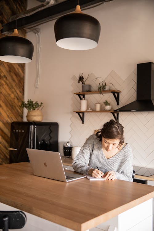 Pensive young businesswoman in gray sweater sitting at table with netbook in modern kitchen and taking notes while working on remote project in home workplace