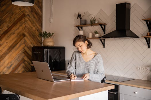 A young woman writes notes while working on a laptop in a stylish kitchen setting.