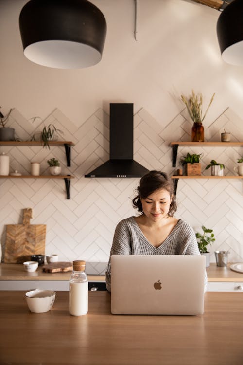 Young female student in gray sweater sitting at wooden desk with laptop and bottle of milk near white bowl during distance education at home