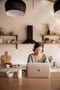 Young female student in gray sweater sitting at wooden desk with laptop and bottle of milk near white bowl during distance education at home