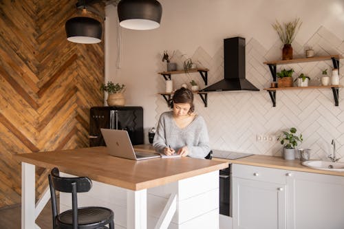 Busy female freelancer with laptop taking notes in kitchen