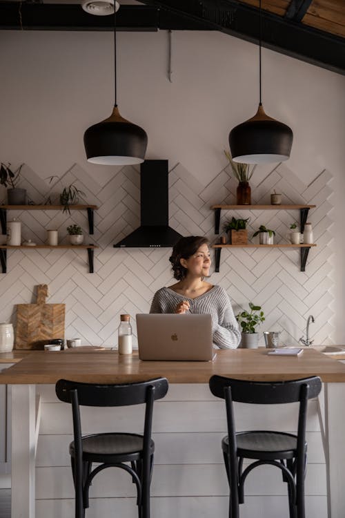 Cheerful female in gray sweater sitting at wooden counter with laptop and bottle of milk while browsing internet on netbook during free time at home and looking away
