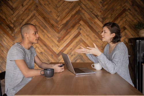 Focused woman explaining opinion to ethnic male coworker during business teamwork sitting at table with laptop and coffee cups in cozy kitchen against wooden wall and looking at each other