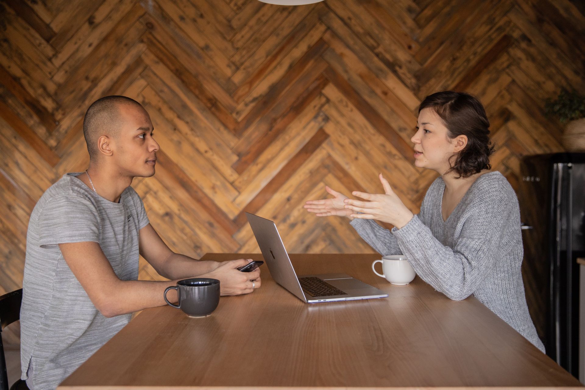 Focused woman explaining opinion to ethnic male coworker during business teamwork sitting at table with laptop and coffee cups in cozy kitchen against wooden wall and looking at each other