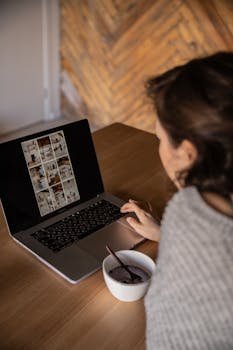 From above unrecognizable female interior designer in gray sweater sitting at wooden table with food in bowl and browsing laptop while working on remote project