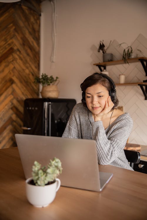 Calm female in casual clothes watching video on laptop while sitting in headphones in cozy kitchen