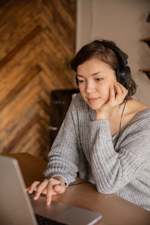 Relaxed female in cozy sweater using laptop while sitting at wooden table in headphones