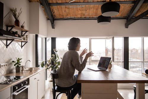 Back view of young female expressively talking via laptop while sitting at wooden table in spacious kitchen