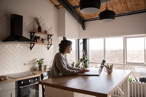 Side view of young female in casual clothes typing message on laptop while sitting at wooden table in kitchen