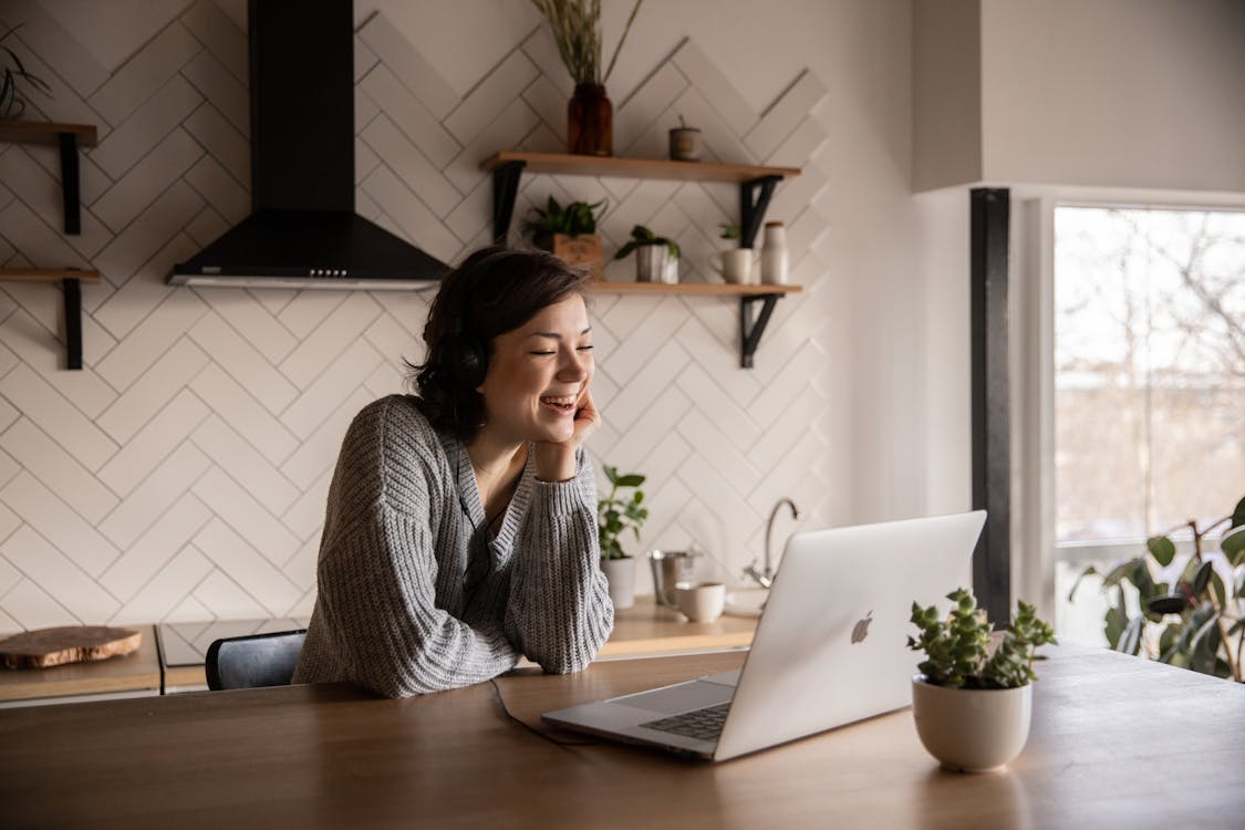 Free Young cheerful female smiling and talking via laptop while sitting at wooden table in cozy kitchen Stock Photo