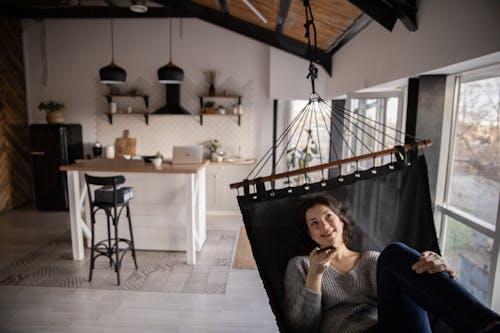 Cheerful female in casual clothes speaking on smartphone via speaker while resting in hammock inside of apartment and looking away