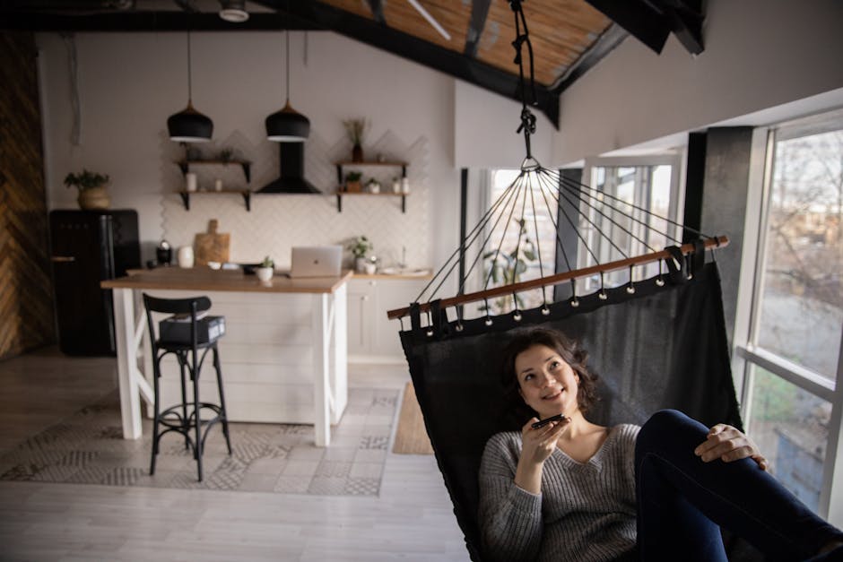 Cheerful female in casual clothes speaking on smartphone via speaker while resting in hammock inside