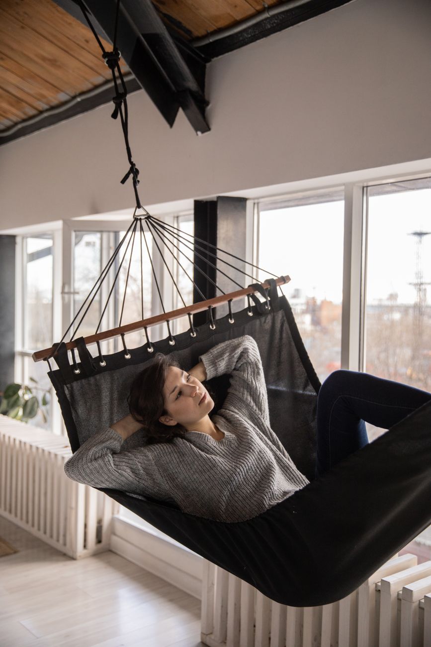 Relaxed pensive female in casual clothes resting in hammock with black canvas hanged below ceiling in apartment